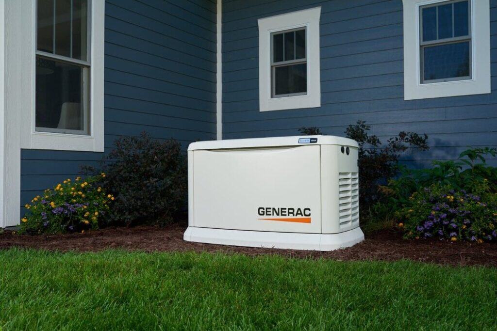 A Generac Generator Is Installed Next To A Blue House With Two Windows. It Sits On A Mulch Bed, Surrounded By Green Grass And Small Flowering Plants.