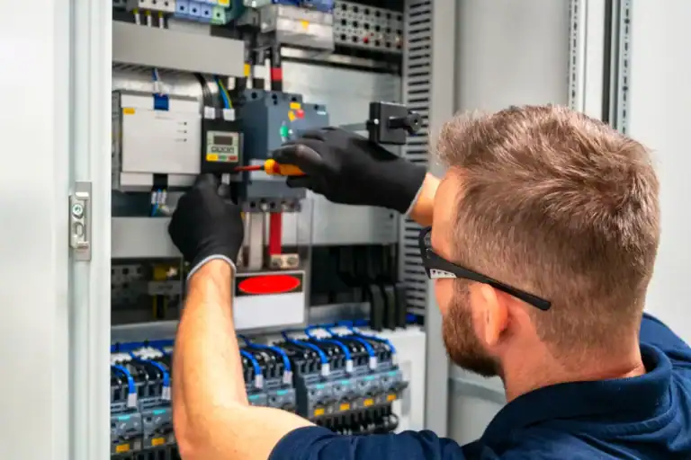 A Technician Wearing Gloves Uses A Screwdriver To Adjust Wiring Inside An Electrical Control Panel.