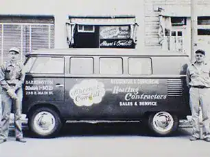 Two Men Stand On Either Side Of A Vintage Van With Signage For A Heating And Contracting Business, Parked In Front Of A Storefront.