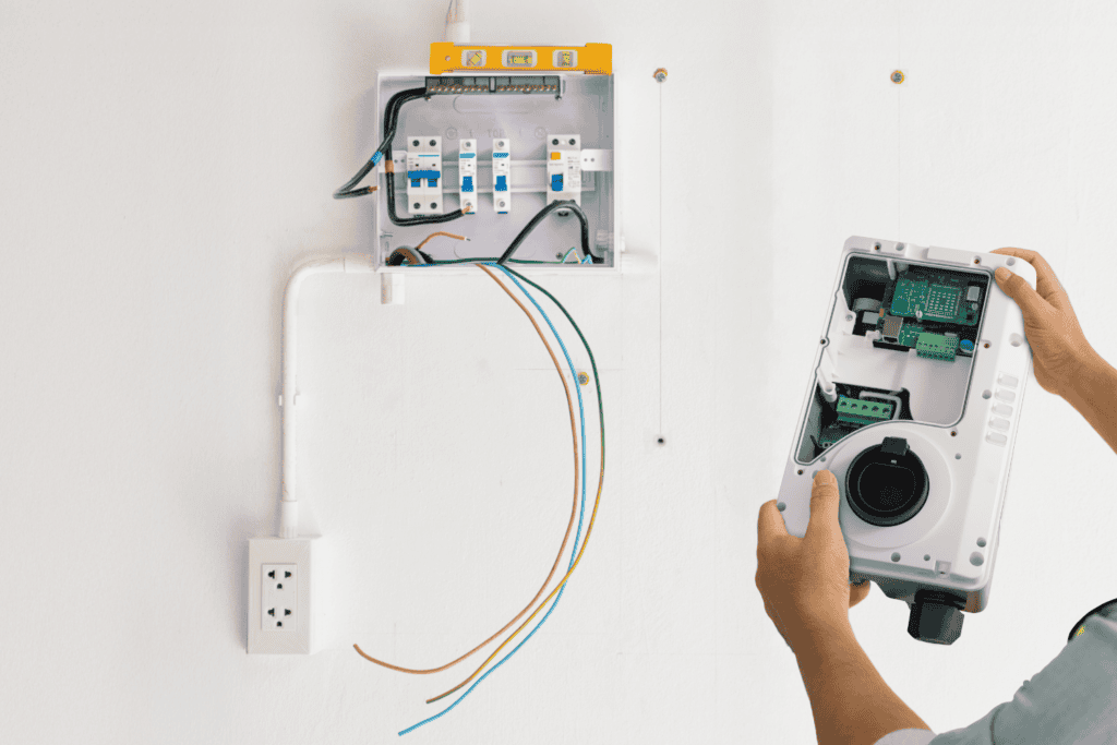 A person installs an electrical device with exposed wiring beside an open circuit breaker panel on a white wall.