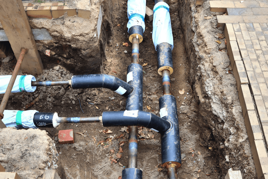 Three insulated pipes, partially wrapped in blue material, run through a trench at a construction site with exposed soil and brickwork visible.
