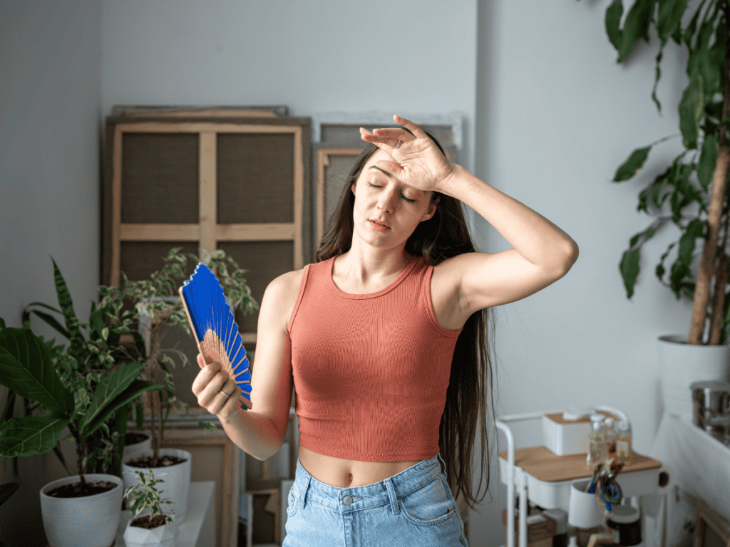 A woman in a sleeveless top and jeans fans herself with a blue fan and wipes her forehead, standing indoors among houseplants.